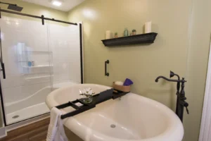 Bright, modern bathroom with sage green walls, featuring a freestanding white soaking tub with a bath tray, a walk-in shower with black trim, and dark shelving decorated with candles.