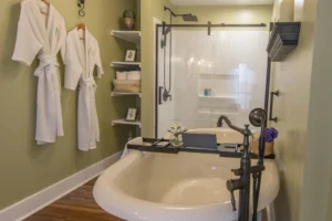Spa-like bathroom with sage green walls, featuring a prominent freestanding soaking tub, a walk-in shower with black framing, corner shelves holding rolled towels, and two plush white robes hanging on the wall.