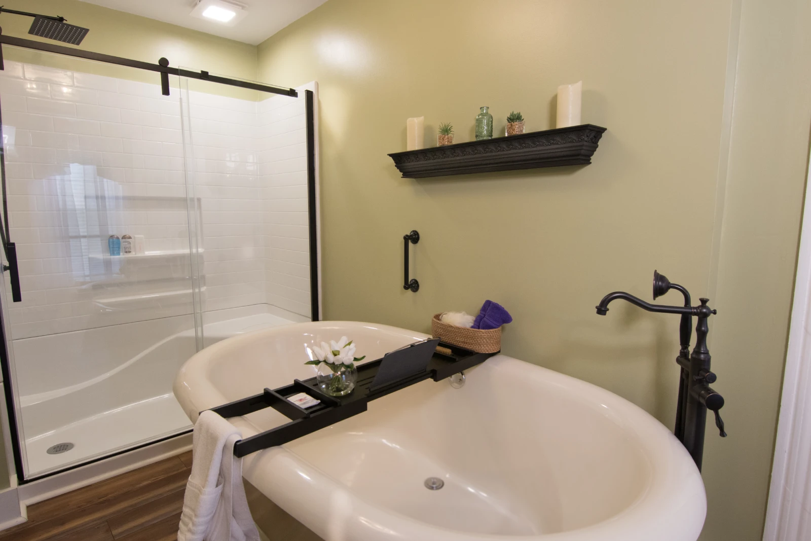 Bright, modern bathroom with sage green walls, featuring a freestanding white soaking tub with a bath tray, a walk-in shower with black trim, and dark shelving decorated with candles.