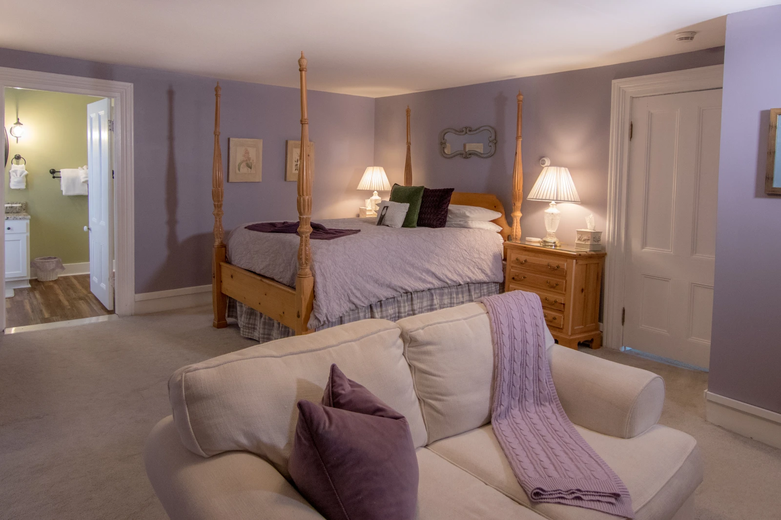 Bedroom suite decorated in lilac and white, featuring a light wood four-poster bed, an off-white sofa in the foreground with purple accents, and an open doorway to a bathroom on the left.