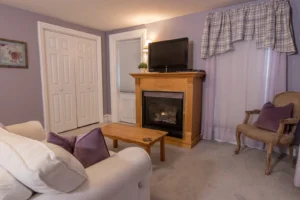 Cozy living area of a suite with lilac walls, featuring a white sofa in the foreground with purple pillows, a gas fireplace with a light wood mantel and a TV above it, and a classical armchair.