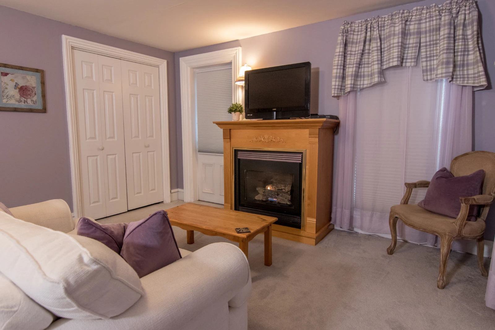 Cozy living area of a suite with lilac walls, featuring a white sofa in the foreground with purple pillows, a gas fireplace with a light wood mantel and a TV above it, and a classical armchair.
