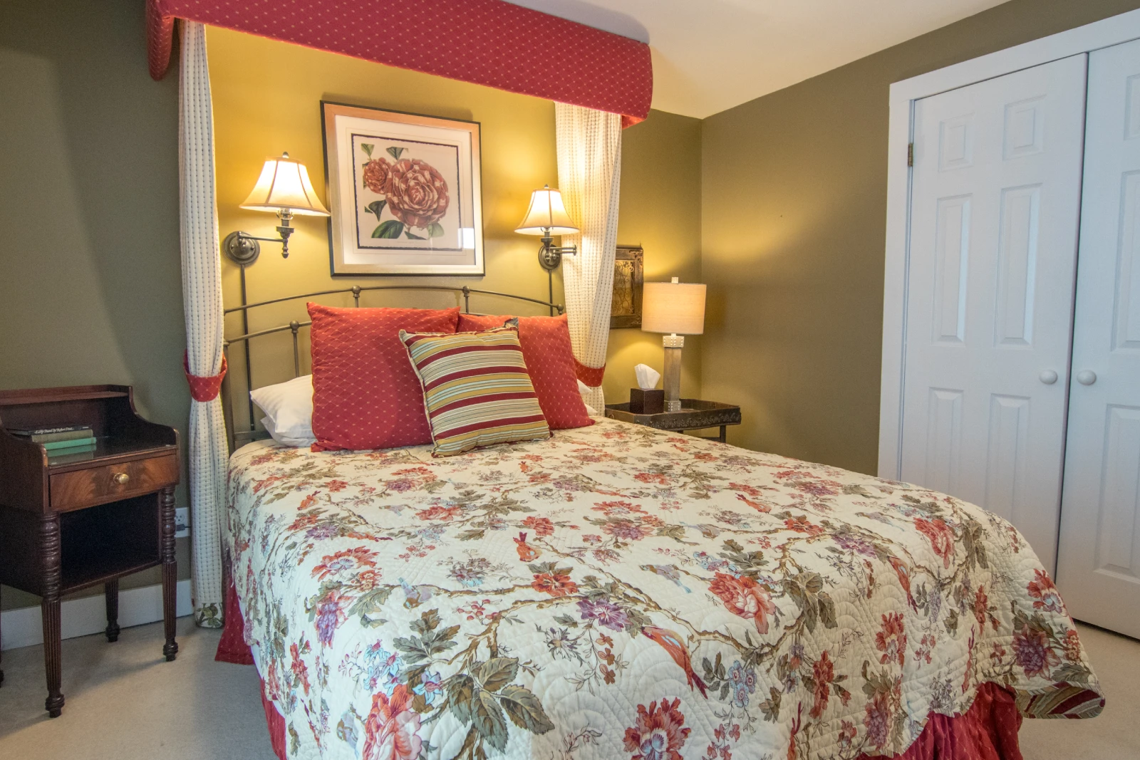 Rose-themed bedroom with olive green walls, featuring a bed with a red floral quilt and a red fabric canopy headboard, and two dark wood nightstands with lamps.