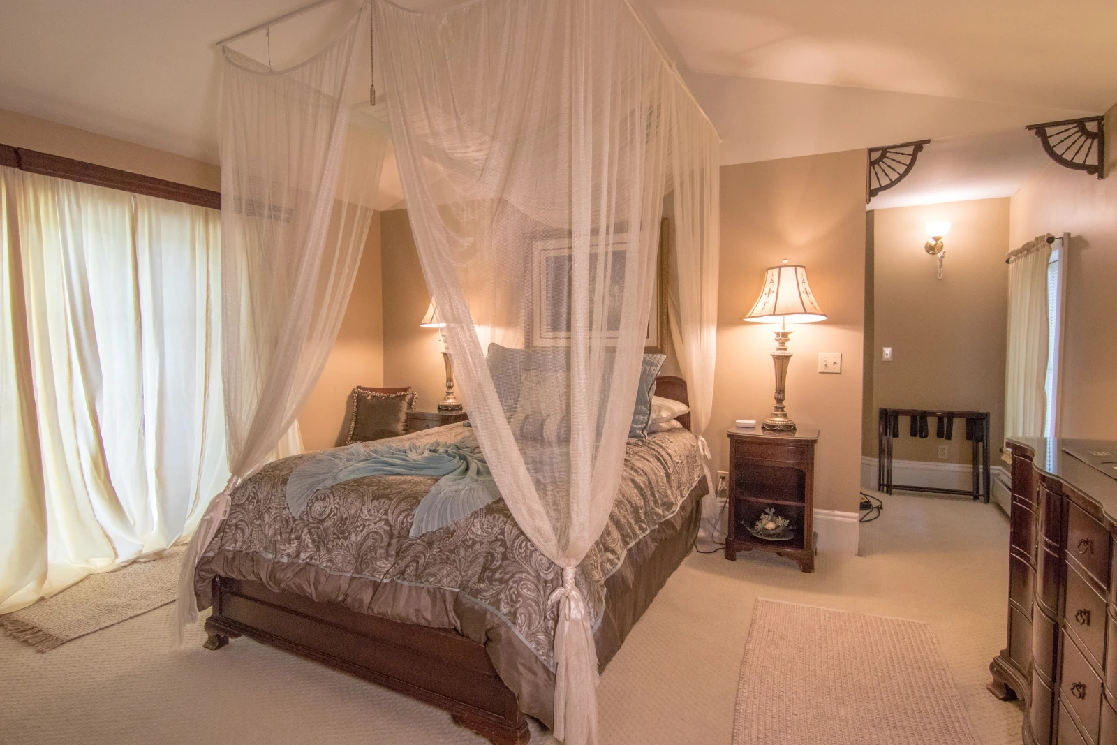 Luxurious bedroom featuring a dark wood bed with a white sheer canopy, gold and light blue patterned bedding, a dark wood nightstand with a lamp, and tan walls.