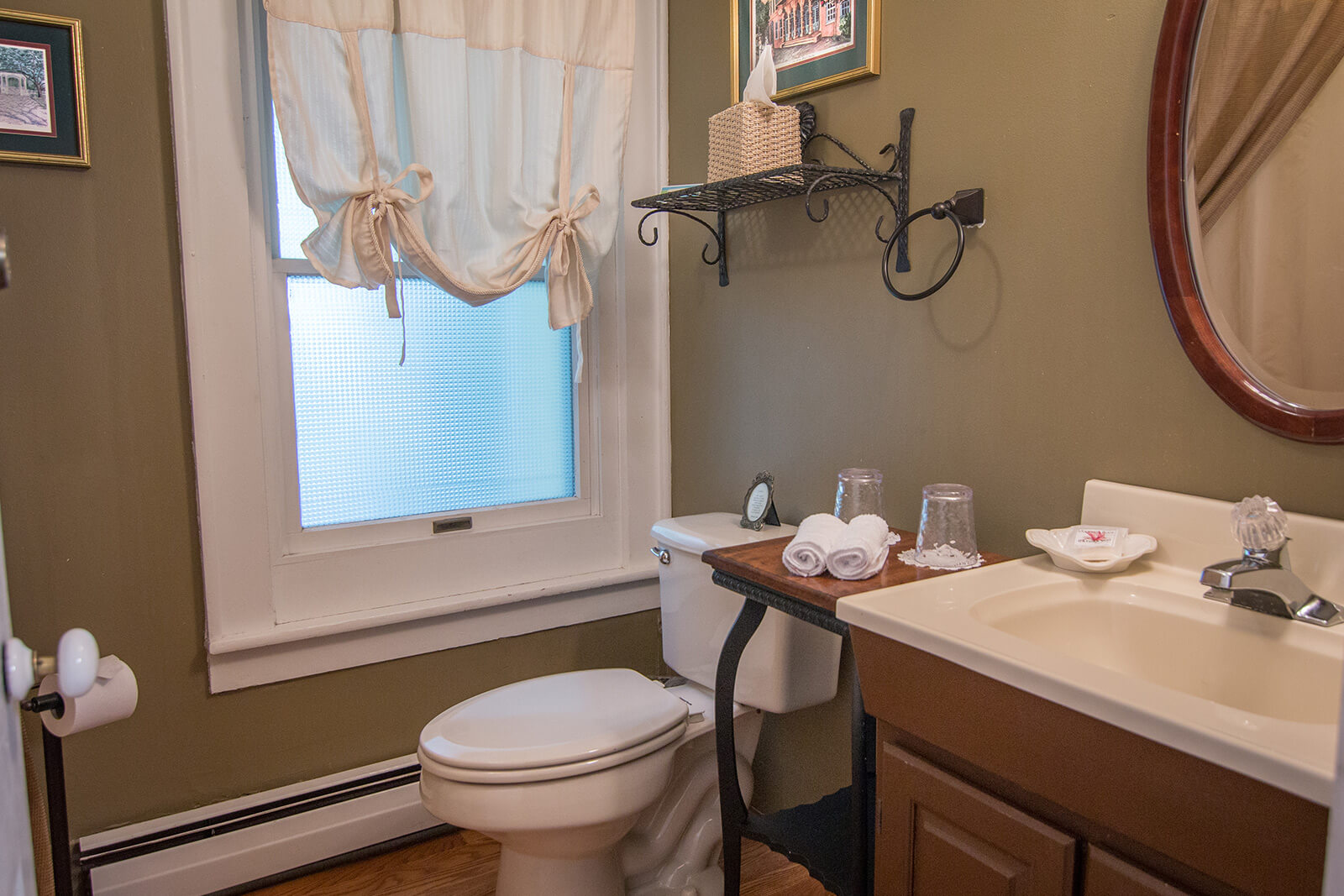 Cozy bathroom with olive green walls, featuring a frosted window with tie-up curtains, a simple vanity sink, and a toilet with a small wooden shelf above it holding fresh towels and glasses.
