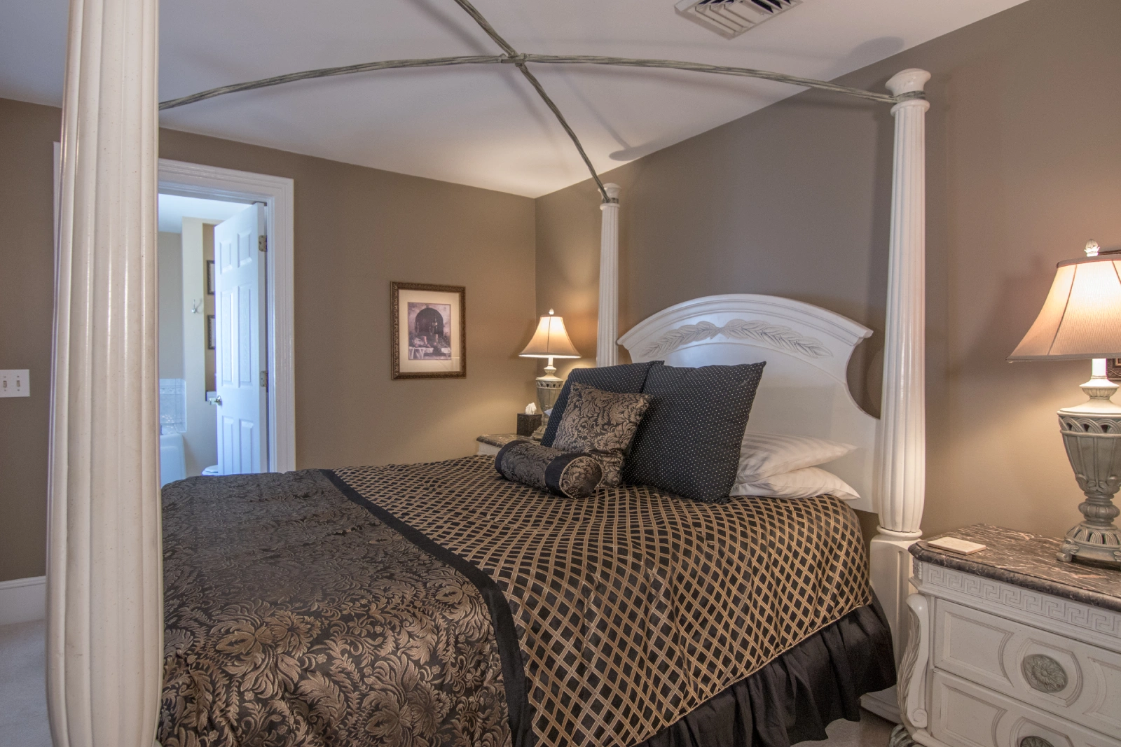 Elegant bedroom with taupe walls, featuring a white, ornate canopy bed (without fabric) dressed in black and gold patterned bedding, and an open door to the bathroom.