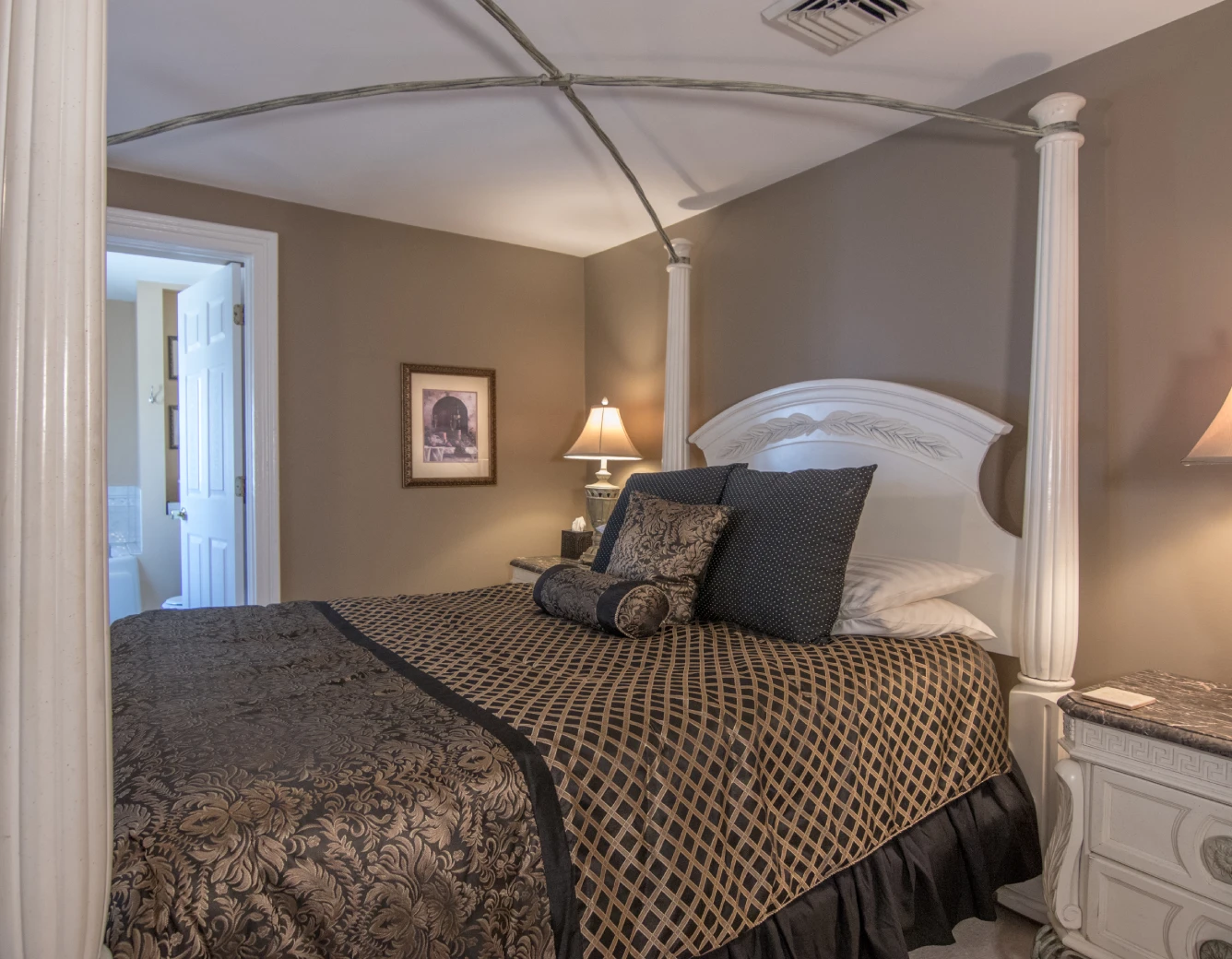 Elegant bedroom with taupe walls, featuring a white, ornate canopy bed (without fabric) dressed in black and gold patterned bedding, and an open door to the bathroom.