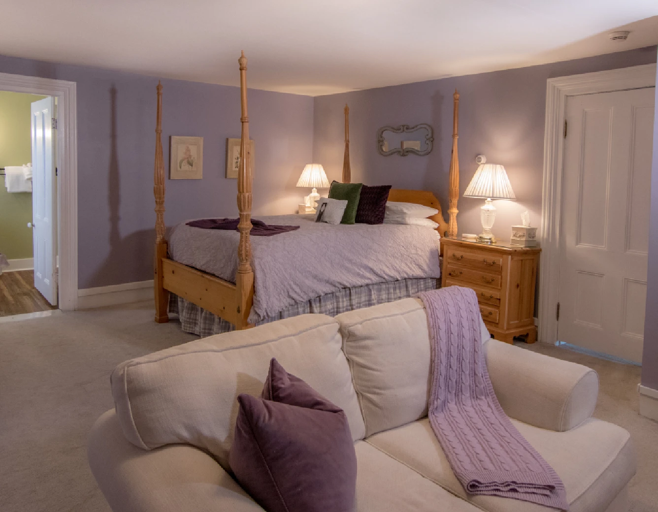 Bedroom suite decorated in lilac and white, featuring a light wood four-poster bed, an off-white sofa in the foreground with purple accents, and an open doorway to a bathroom on the left.