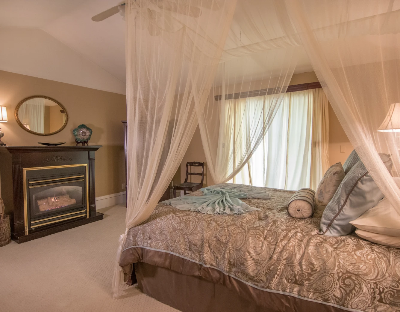 Elegant bedroom with a large bed draped in canopy netting and gold patterned bedding, a dark wood fireplace with a round mirror above it, and bright curtains behind the bed.